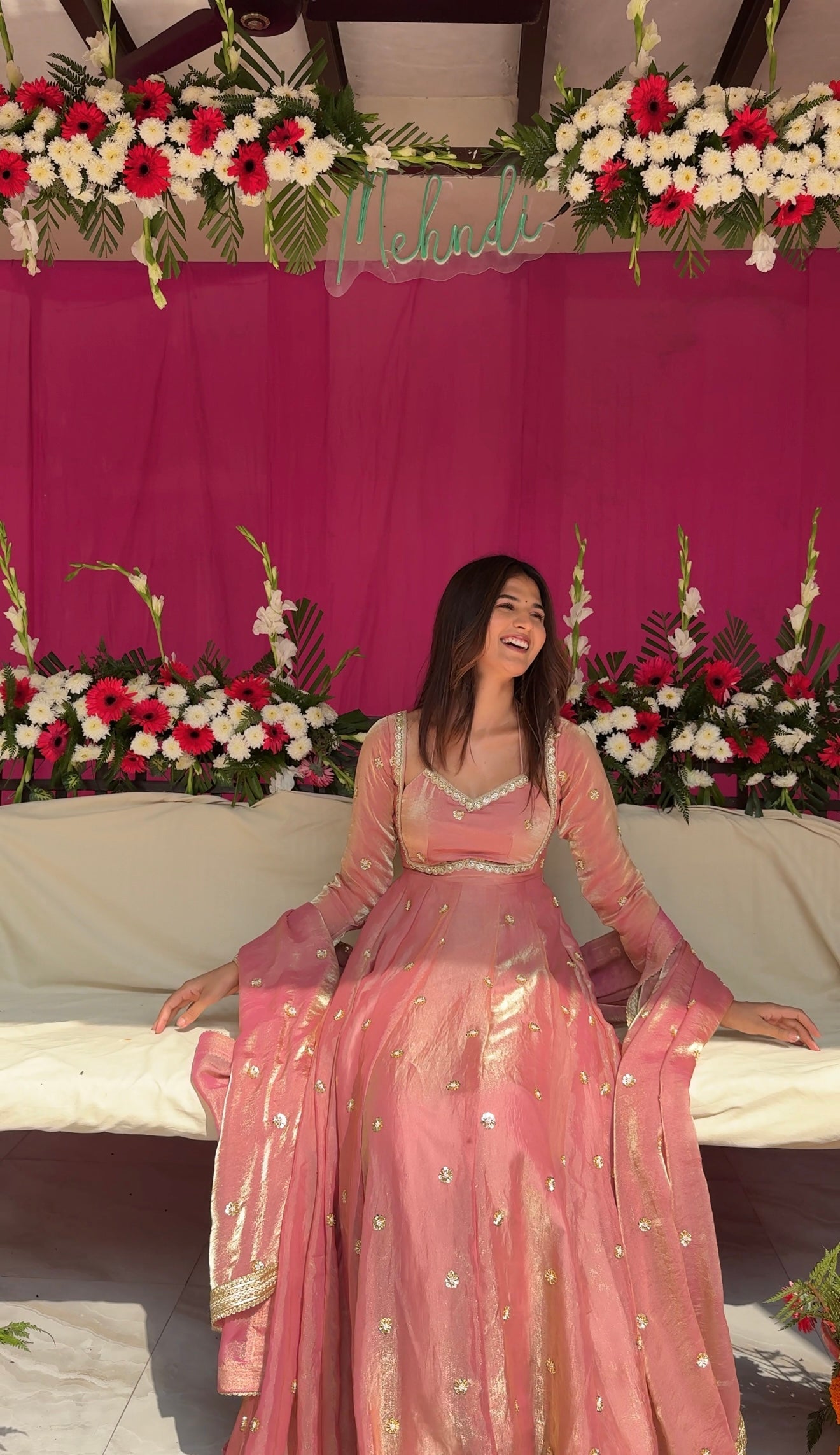 Woman in a pink traditional outfit sitting on a decorated stage with flowers and a pink curtain.