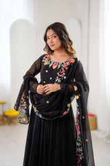 A woman modeling a black traditional outfit with vibrant floral embroidery in pink, yellow, and green, paired with a matching sheer black dupatta draped over her shoulders.