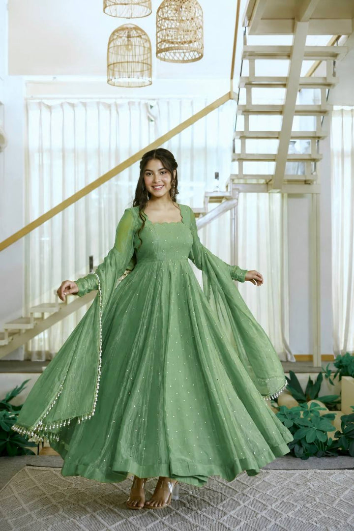 A woman wearing a flowing sage green Anarkali gown with sheer long sleeves and a matching dupatta, standing in a bright modern interior with a staircase and hanging lanterns.