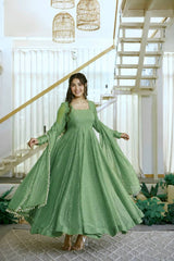 A woman wearing a flowing sage green Anarkali gown with sheer long sleeves and a matching dupatta, standing in a bright modern interior with a staircase and hanging lanterns.