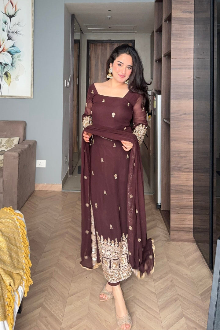 Woman in a dark red traditional outfit standing in a room with wooden flooring and a gray wall.