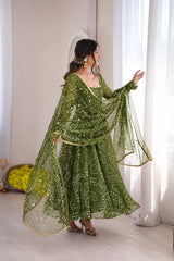 A woman wearing a green embroidered traditional dress with a matching sheer dupatta, posing in a bright room with white curtains.