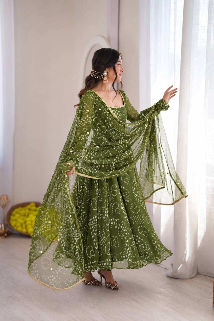 A woman wearing a green embroidered traditional dress with a matching sheer dupatta, posing in a bright room with white curtains.