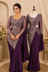 A woman and a mannequin display matching purple traditional garments with ornate silver embroidery. The woman wears a draped sari-style dress, while the mannequin shows a similar outfit on a dress form in an indoor styled setting with neutral walls, a vase of flowers, and a small table.