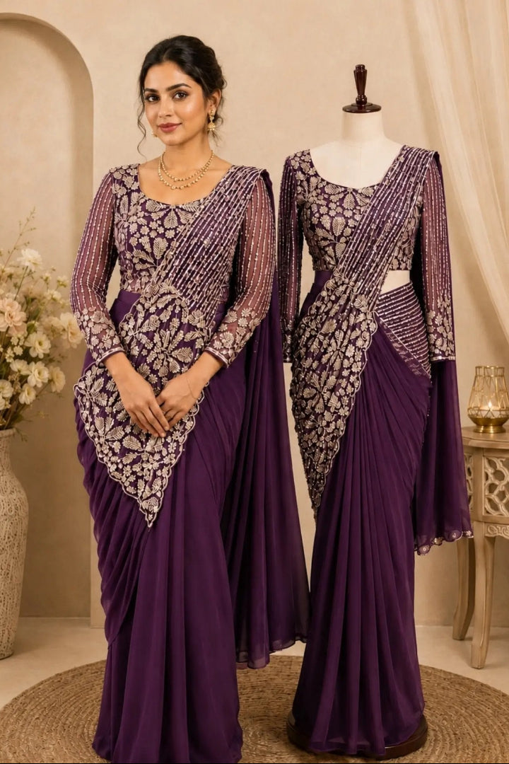 A woman and a mannequin display matching purple traditional garments with ornate silver embroidery. The woman wears a draped sari-style dress, while the mannequin shows a similar outfit on a dress form in an indoor styled setting with neutral walls, a vase of flowers, and a small table.