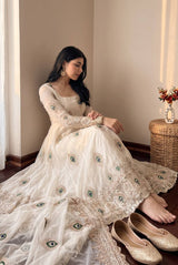 A woman sits barefoot on a wooden floor wearing a long cream/off-white embroidered gown with peacock-eye motif details and matching sheer dupatta. She is styled in a warm indoor setting with a woven side table and gold flat shoes nearby.