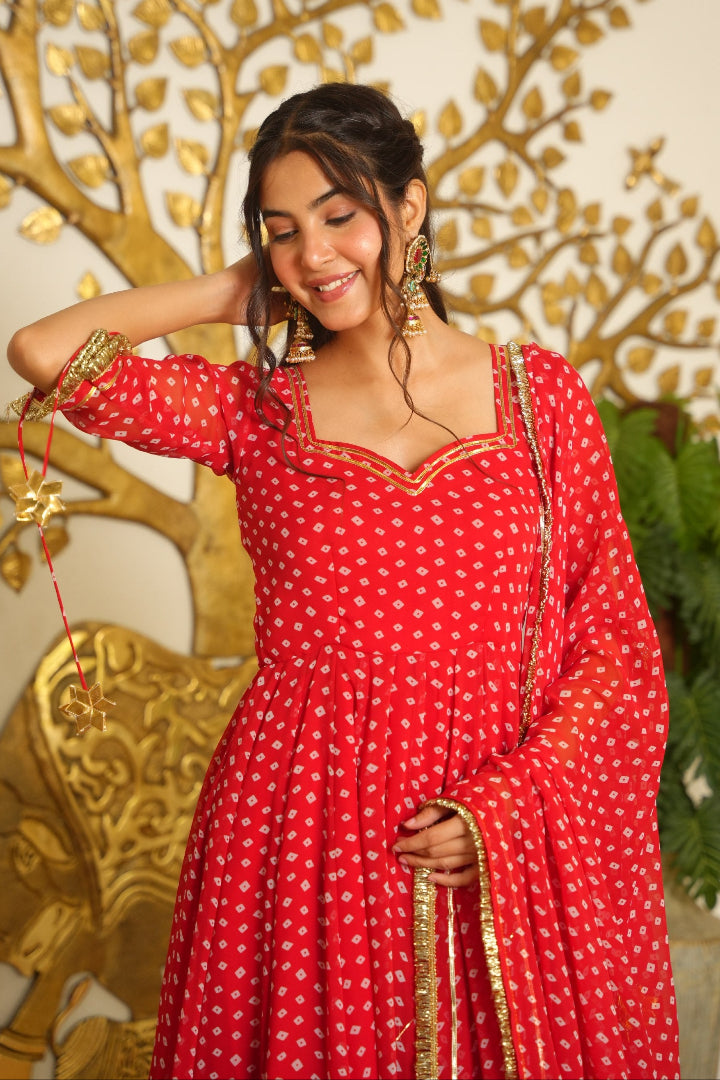 A woman wearing a red traditional dress with gold patterns and a sheer dupatta, standing in front of a decorative gold tree wall and green plant.