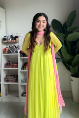 Woman in a bright yellow dress with pink dupatta standing indoors next to a shelf and plant.