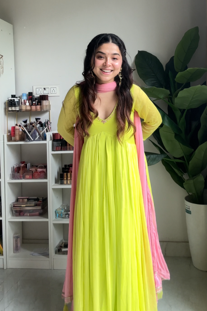 Woman in a bright yellow dress with pink dupatta standing indoors next to a shelf and plant.