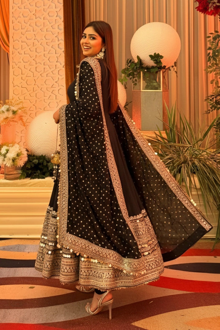 Woman in a black and gold traditional outfit standing in a decorated room with plants and decorative items.