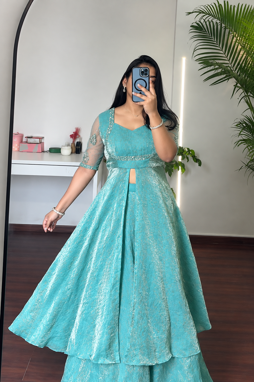 A woman takes a mirror selfie wearing a flowing turquoise blue anarkali-style gown with sheer embroidered sleeves and a flared skirt, photographed indoors in a room with a plant and white wall.