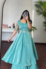 A woman takes a mirror selfie wearing a flowing turquoise blue anarkali-style gown with sheer embroidered sleeves and a flared skirt, photographed indoors in a room with a plant and white wall.