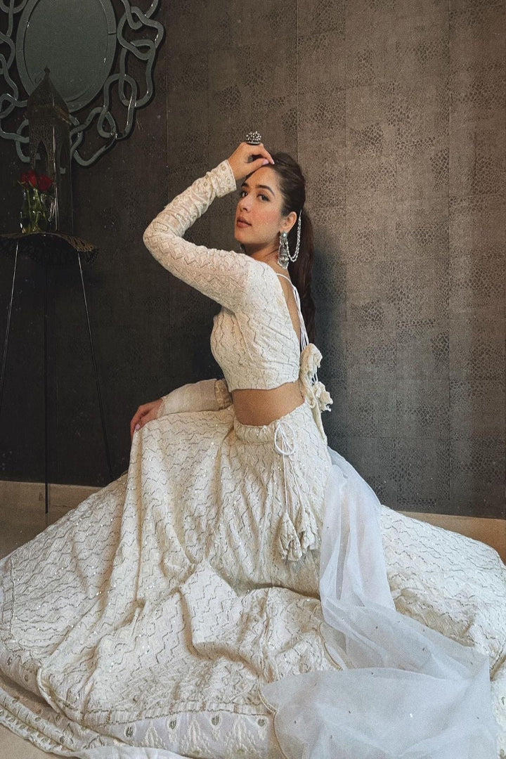 Woman in a white embroidered traditional outfit sitting against a textured wall.
