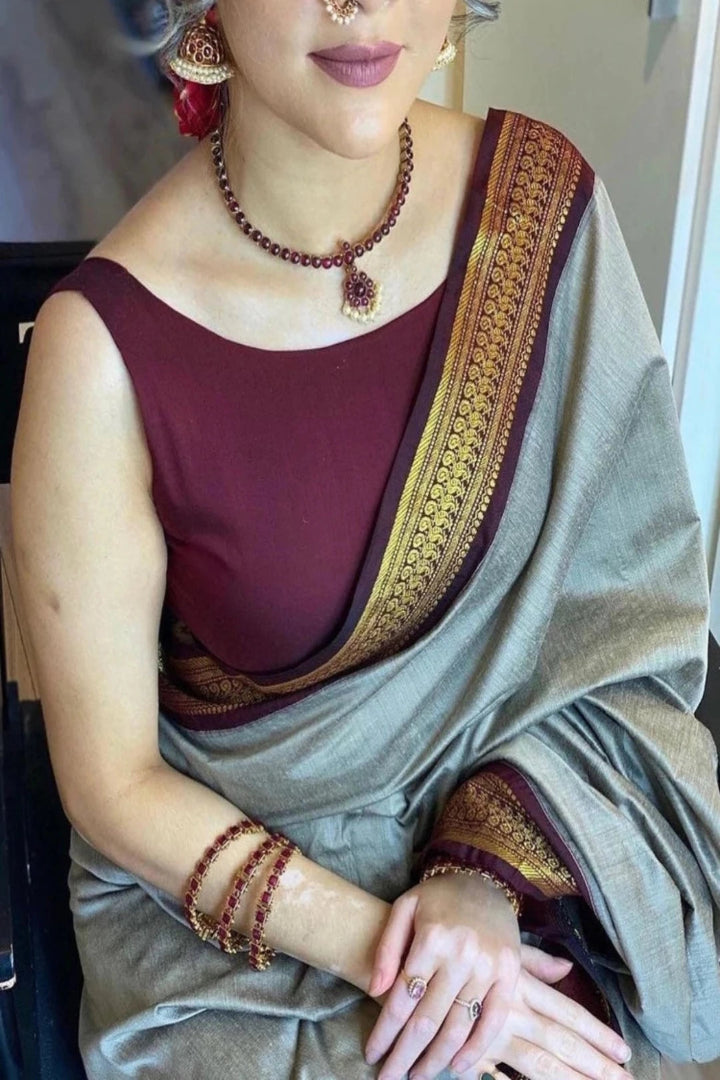 Woman wearing a maroon blouse with gold border and a light gray saree, accessorized with jewelry.