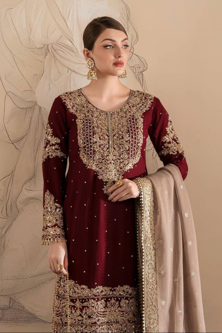 Woman wearing a maroon and gold embroidered traditional outfit with a beige dupatta against a beige background.
