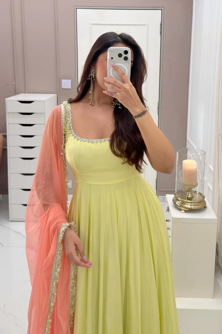 A woman takes a mirror selfie wearing a light yellow sleeveless gown with a peach dupatta featuring gold embroidery, in a modern room with beige walls and white furniture.