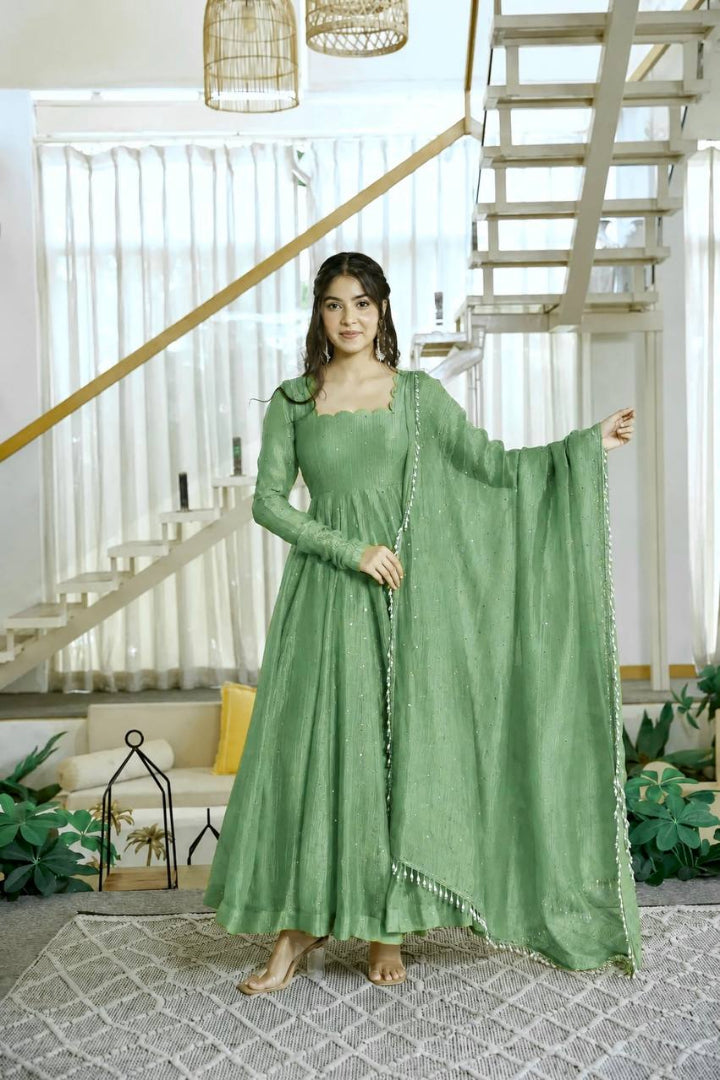 A woman stands barefoot in a bright indoor setting wearing a long sage green anarkali-style dress with a matching dupatta draped over one arm. The room has white curtains, a staircase, pendant lights, and plants.