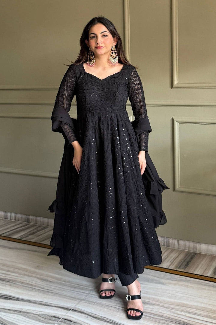 Woman wearing a black dress with intricate detailing in a room with a neutral-colored wall.