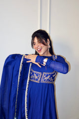 A woman wearing a royal blue traditional outfit with gold embroidery and a matching dupatta, posing against a plain white wall.