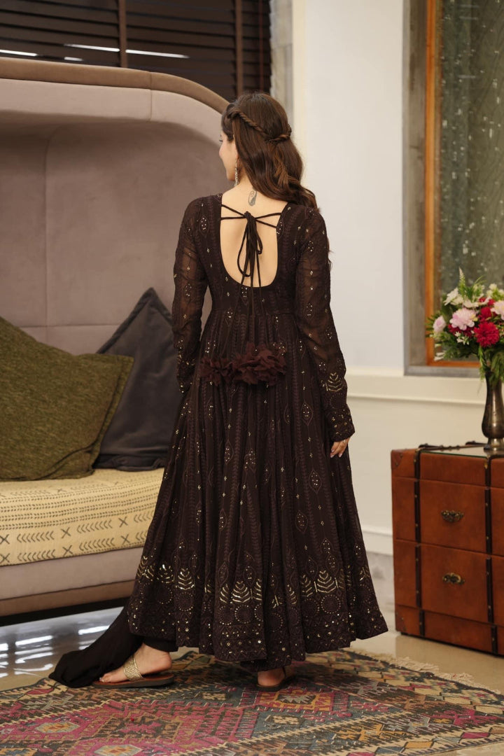 Woman in a long, dark brown lace dress standing in a room with a couch and decorative items.
