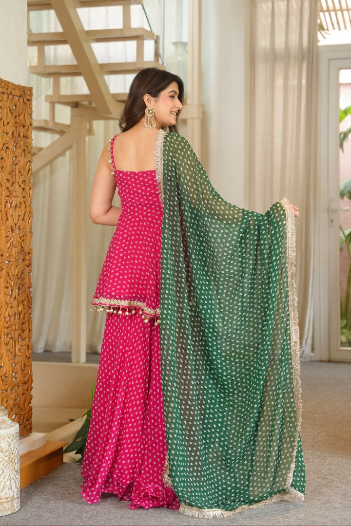 A woman wearing a bright pink sleeveless traditional dress with a matching green dupatta featuring white printed motifs, standing in an indoor setting with a staircase and carved wooden screen in the background.
