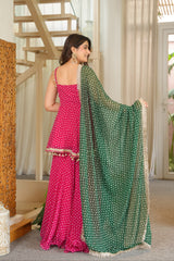 A woman wearing a bright pink sleeveless traditional dress with a matching green dupatta featuring white printed motifs, standing in an indoor setting with a staircase and carved wooden screen in the background.