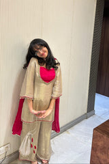 A woman wearing a traditional Indian outfit consisting of a beige kurta with a bright pink inner lining and matching beige trousers, standing in an indoor hallway.