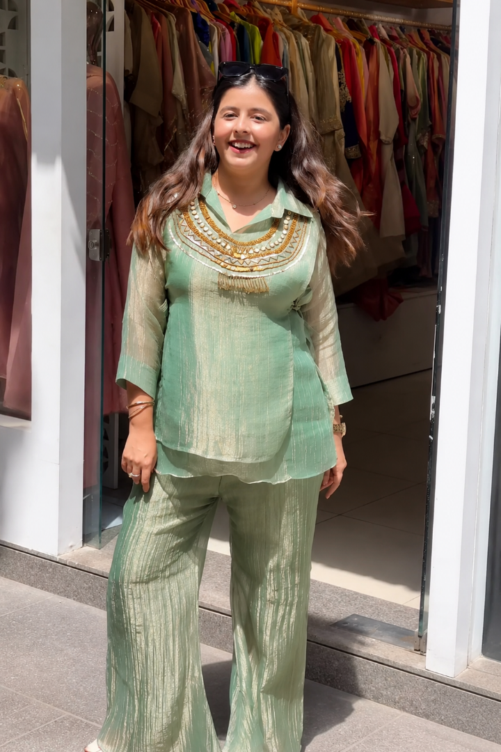 A woman wearing a light sage green two-piece set with gold embroidery on the neckline and matching pleated trousers, standing in front of a clothing store doorway.