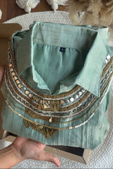 A sage green linen shirt with intricate gold and silver bohemian embroidery, sequins, and coin details, held in a hand over a cardboard box.