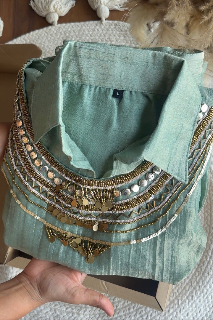 A sage green linen shirt with intricate gold and silver bohemian embroidery, sequins, and coin details, held in a hand over a cardboard box.