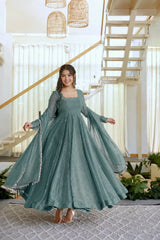 A woman wearing a flowing sage green anarkali gown with sheer sleeves and silver embellishments, posing in a bright, modern indoor setting with a staircase and plants.
