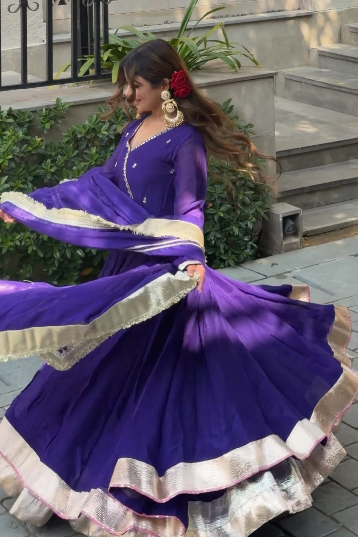 Woman in a purple traditional outfit with white and gold details, standing outdoors.