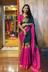 Woman in a green and pink saree standing in a room with colorful clothes in the background.