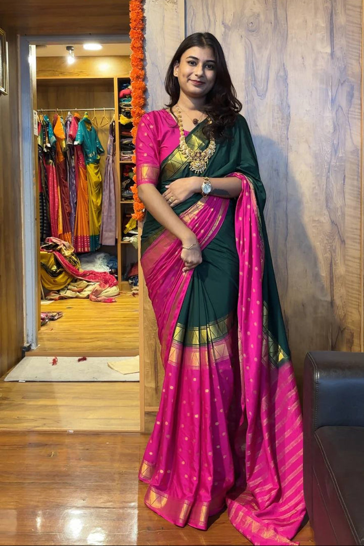 Woman in a green and pink saree standing in a room with colorful clothes in the background.