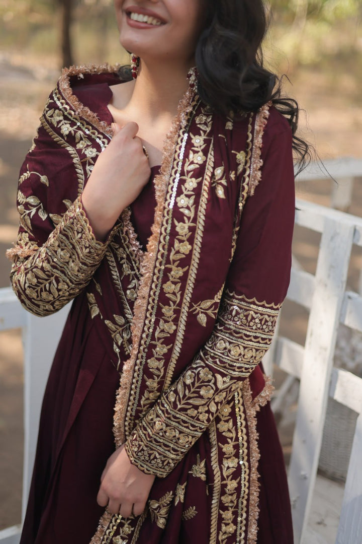 Woman wearing a maroon traditional outfit with gold embroidery, standing outdoors.