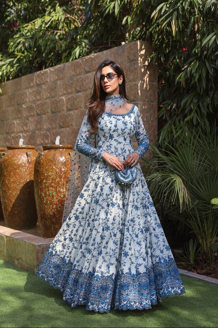 Woman in a blue floral dress standing outdoors with greenery and stone wall in the background