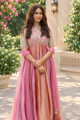 A Mrunal Thakur wearing a traditional pink and peach embroidered Anarkali gown stands outdoors in a garden courtyard with stone walls and potted plants.