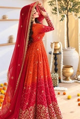 A woman modeling a traditional Indian anarkali lehenga choli in a vibrant red and orange gradient, adorned with intricate gold embroidery and a matching red dupatta with gold borders, set against a decorated indoor background.