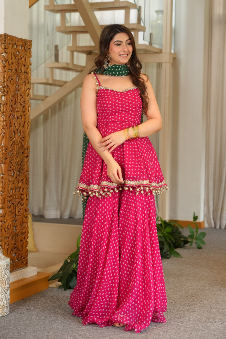 A woman models a vibrant pink and white polka dot traditional Indian outfit consisting of a sleeveless top, flared pants, and a green patterned dupatta in an indoor setting with a staircase and curtains.