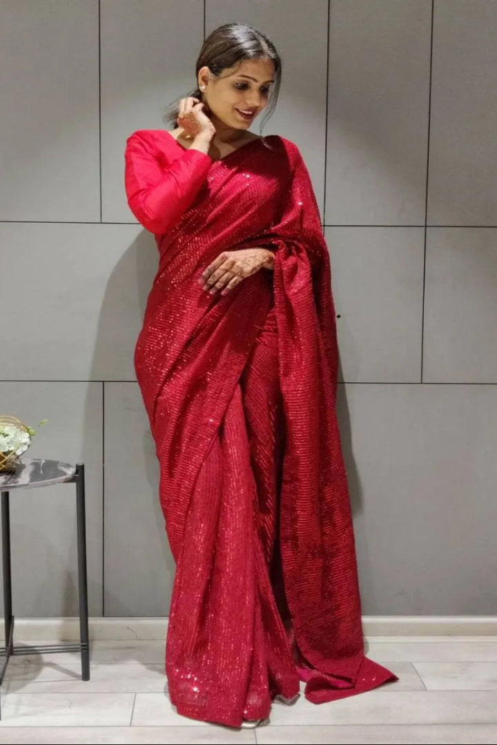 Woman in a red saree standing against a gray wall with a small table and flowers in the background.