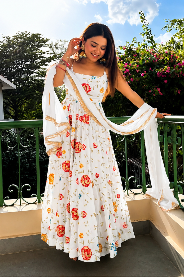 A woman stands on a balcony wearing a white Anarkali gown decorated with red, orange, and yellow floral prints, holding a matching white dupatta with gold border details.