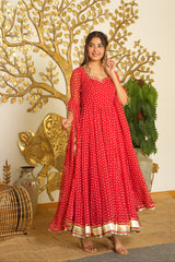 A woman models a red floor-length anarkali dress with white polka dots and a gold border, standing in front of a decorative gold tree wall piece.