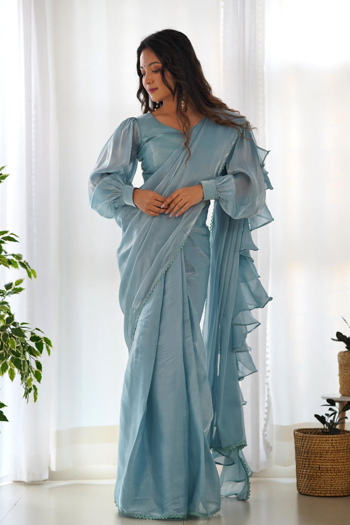 Woman wearing a light blue saree standing in a room with white curtains and a plant.