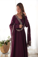Woman wearing a long, dark purple dress with intricate patterns in a bright room.