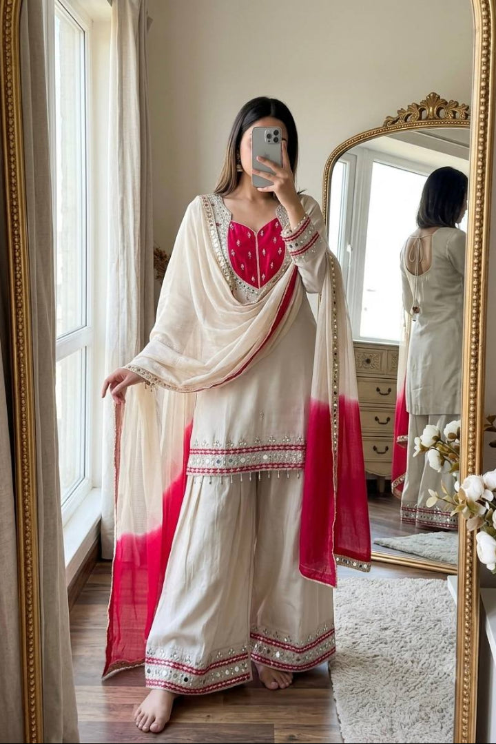 A woman takes a mirror selfie wearing a traditional Indian salwar kameez outfit. The outfit consists of an off-white/cream kurta and wide-leg pants with red and gold embroidery, paired with a matching off-white dupatta featuring a red gradient at the bottom.