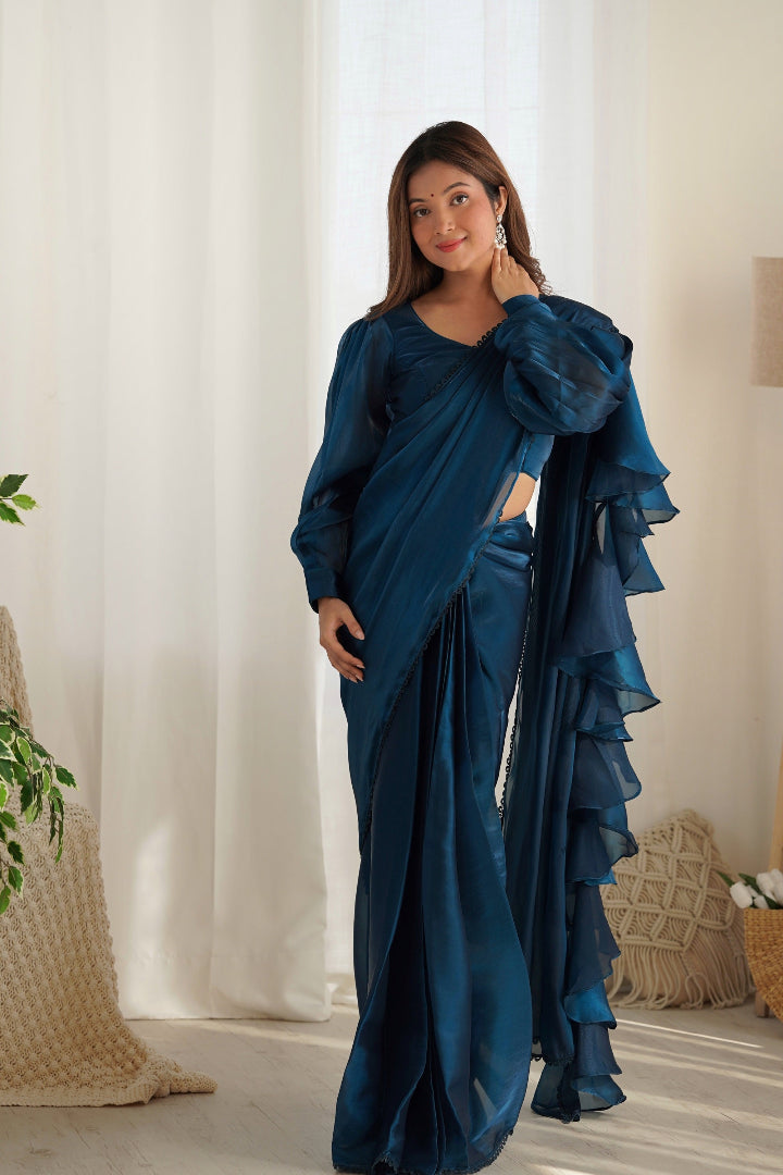 Woman wearing a blue saree with ruffles in a room with white curtains and decorative pillows.
