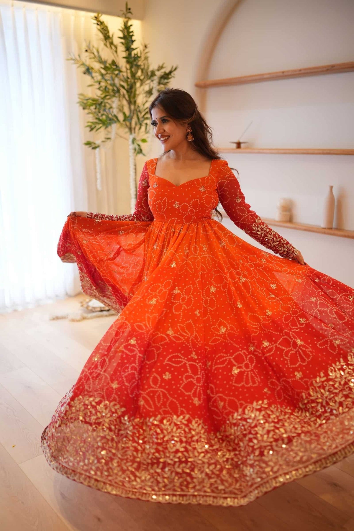 A woman wearing a bright orange-red embroidered traditional gown with long sleeves and a flared skirt, posing indoors in a softly lit room with light wood floors and shelves.
