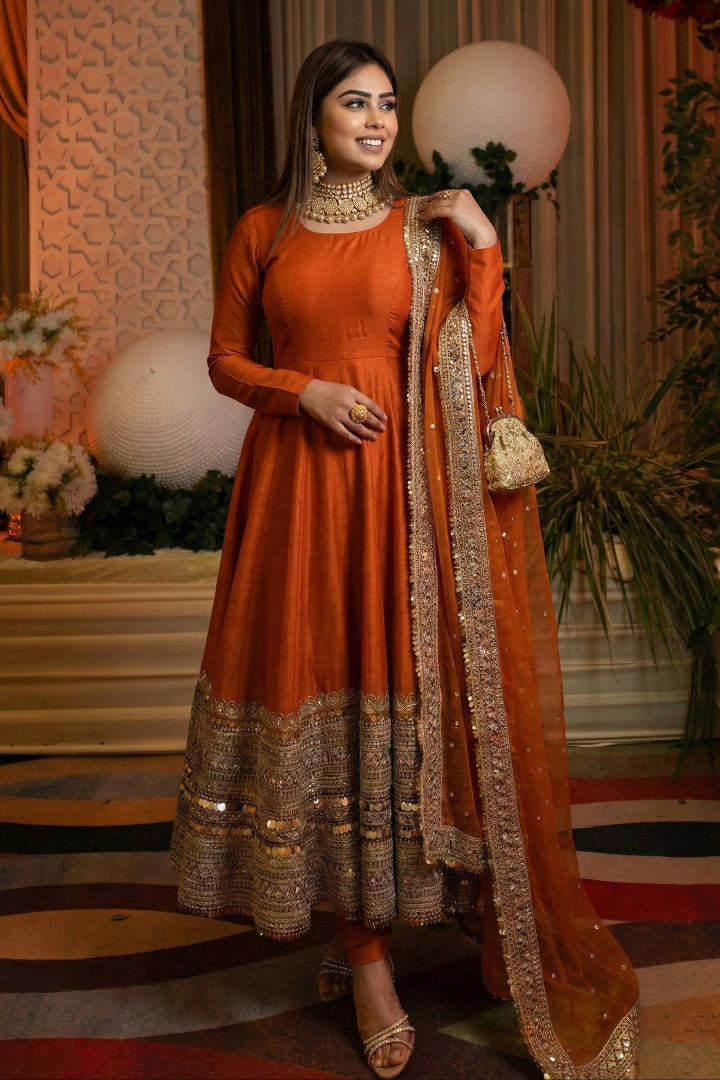 Woman in an orange traditional outfit with intricate designs, standing in a decorated indoor setting.