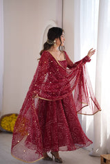 A woman wearing a deep red embroidered traditional outfit with a matching sheer dupatta, standing in a bright room with white curtains.