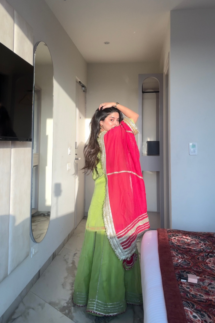 Woman in a green traditional outfit with a pink dupatta standing in a room.